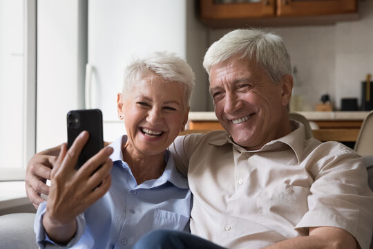Happy joyful senior couple taking home selfie on mobile phone, posing for photo with toothy smile, resting on couch with digital gadget, hugging, having fun, laughing, making video call on smartphone
