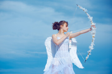 Teen girl in white angels dress, angel cupid valentin shoot with bow arrow. Teenager with feathers...