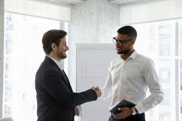 Two diverse smiling business men shaking hands in office boardroom. Employee hiring candidate after job interview. Client thanking manager, lawyer with handshake for consultation, negotiation