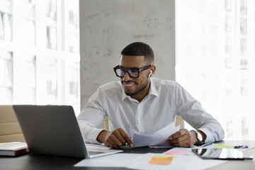 Happy young Afro American business professional man in glasses, formalwear talking on video call in office, negotiating on paper report, using laptop, wireless earbud for communication