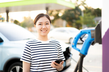 Happy Asian young woman using ev charging application on smartphone connect with the charging...
