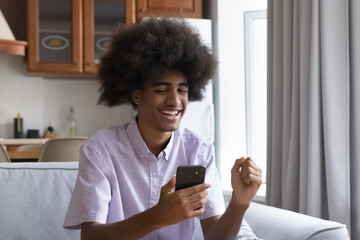 Fototapeta na wymiar Cheerful happy teen African guy with curly fuzzy hair excited with good news in text message, reading notice on smartphone screen, making winner success yes hand gesture on home sofa