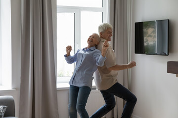 Happy mature active couple dancing to music together in living room, enjoying date, anniversary,...