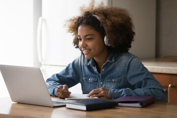 Happy gen Z Black teenage student girl chatting online with teacher, using headphones and laptop at home, writing notes, preparing for school test, college exam, watching learning webinar