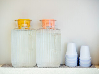 Two plastic bottles with yellow and pink pastel lids full of cold water were placed near the stack of white plastic cups on the shelf to serve customers who come to the cafe for free.