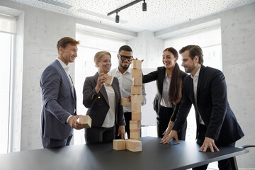 Happy successful diverse business team of different ages building blocked tower on office table together, having fun, laughing, working on teambuilding, unity, cooperation skills, celebrating success
