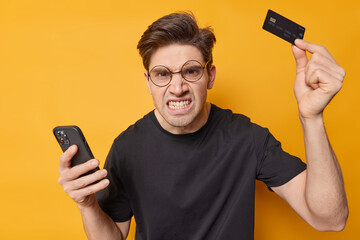 Annoyed man clenches teeth holds mobile phone and credit card for fast online shopping orders...