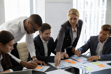 Focused marketing business group working with paper reports and sticky notes together, brainstorming, discussing project. Mature business leader, coach, mentor using scrum method for analysis