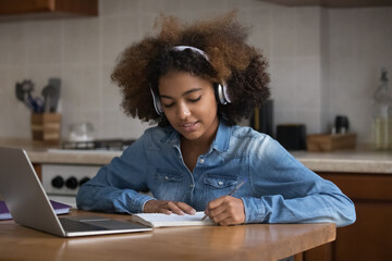 Focused fuzzy haired Black teenage girl studying from home. Gen Z school student, pupil in headphones watching learning webinar on laptop, writing notes, preparing for college test, exam