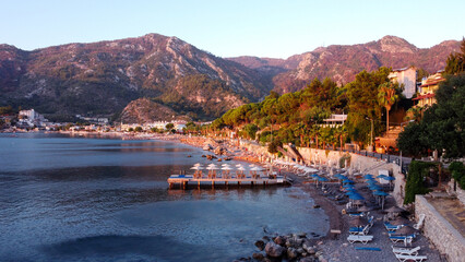 Fototapeta premium An aerial image of a beach in Turunc, Turkey. Sunrise over resort village photografed by drone. Umbrellas and sunbed in row on beach. Turunc, Turkey - September 6, 2021