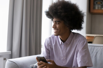Happy teenage fuzzy haired African guy holding digital gadget, looking at window away, smiling at good thoughts. Hipster teen smartphone user sitting on couch. Home portrait