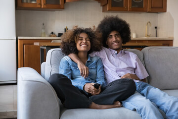 Happy joyful teen African couple watching TV, comedy movie, sitcom series, resting on sofa in...