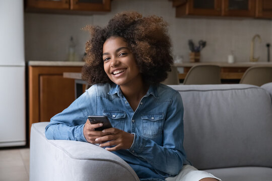 Happy beautiful gen Z teenage girl using cellphone, holding gadget, sitting on couch, looking at camera with cheerful toothy smile. Online app, digital device usage concept. Home portrait
