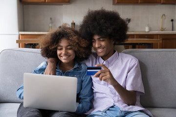 Happy African gen Z teen student couple hugging on couch, enjoying online shopping from home, paying by credit card, making payments for purchase from virtual account, using laptop computer