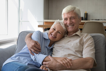Happy grey haired mature couple home candid portrait. Positive smiling senior husband and wife resting on couch at home, hugging with love, care, tenderness, looking away, smiling, laughing