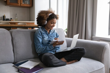 Happy Black teenage student girl in headphones studying foreign language at home, listening learning webinar, audio lecture, course, book on laptop, writing notes, working on high school class project