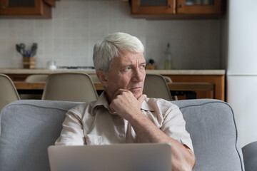 Pensive stressed mature 70s man holding laptop computer, looking away in deep sad thoughts,...