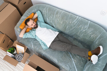 Top view of adult man with boxes moving in new house, lying and relaxing on sofa.