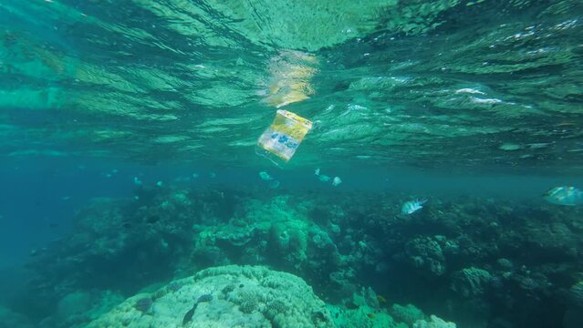 Slow motion, discarded plastic packaging slowly drifting under the surface of water nearby coral reef a tropical fish swimming around. Yellow plastic bag floats in Red Sea