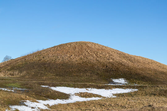 Burial mound covered with yellow grass in culture landscape. Wintertime with patches of snow on the ground.