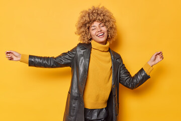 Indoor shot of happy woman shakes arms dances carefree keeps eyes closed smiles toothily wears sweater and leather black coat isolated over yellow background. People emotions feelings concept