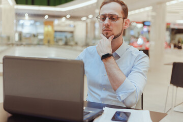 Pensive young Caucasian man sit at table food court work online on laptop pondering thinking