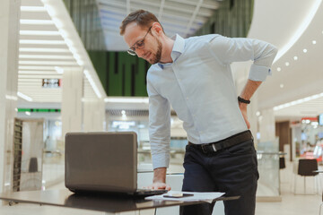 Man holds on to his back, suffers from lower back pain, standing near a desktop with a computer....