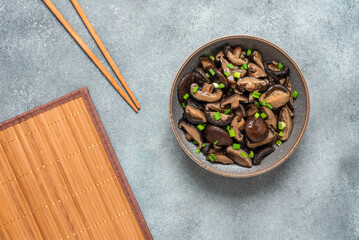 Fried shiitake mushrooms with green onions in a bowl on a bamboo mat. Gray rustic background. Asian vegan food. Top view.