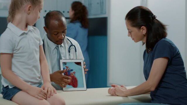 Doctor showing heart image on tablet to woman and kid, explaining cardiology diagnosis and cardiovascular examination at appointment. Pediatrician holding device at consultation.