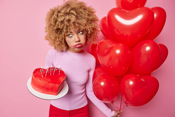 Upset lonely sorrowful woman hates Valentines Day feels unhappy has leaked makeup after crying holds delicious cake and heart shaped balloons isolated over pink background. Holidays concept.
