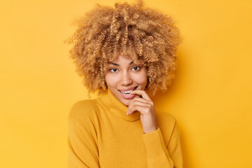 Flirty charming lovely woman bites finger looks directly at camera ha fair curly bushy hair wears casual jumper isolated over vivid yellow background. People facial expressions and dreams concept
