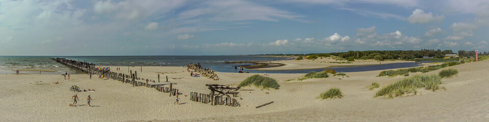 Panoramic photo of the mouth of the "Sventoji" river. You can see the old ruined bridge, the sandy beach, the sea, the sky with clouds. Stone embankment. Resting people.
