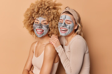 Indoor shot of happy young pretty women apply clay masks smile broadly dressed casually stand closely to each other pose against brown background. Facial treatments beauty and skin care concept