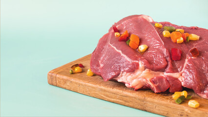 A piece of raw meat with pieces of frozen vegetables on a wooden backing blue background