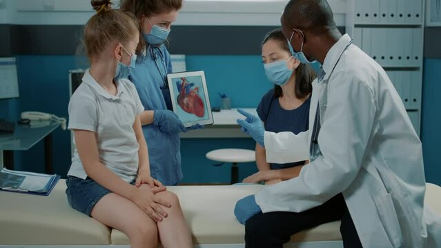 Nurse holding cardiology picture and medic explaining heart condition at examination. Assistant and doctor analyzing cardiovascular diagnosis with child and mother during covid 19 pandemic.