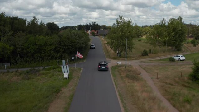 Forwards tracking of car passing through village. Danish flags on poles along street. Horses grazing on fresh green grass. Denmark