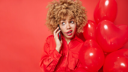 Unhappy crying woman answers phone call upset nobody congratulated her with Valentines Day wears stylish shirt holds heart shaped helium balloons isolated over red background with copy space