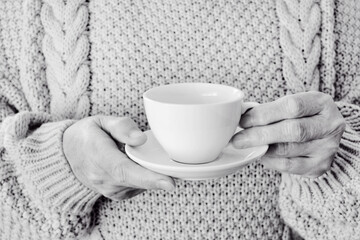 Elderly hands of a woman hold a white cup and saucer with tea at home close-up against the background of a knitted sweater
