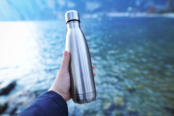 A man's hand holds a water bottle close up against the background of clear water of a lake with a turquoise hue with the shore in the distance. Concept of shortage of fresh water supplies. Background.