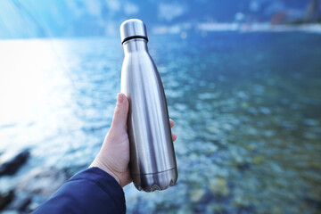 A man's hand holds a water bottle close up against the background of clear water of a lake with a...