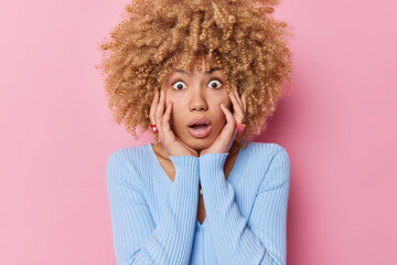 Shocked curly haired woman keeps hands on cheeks stares bugged eyes at camera has surprised exression holds breath wears casual blue jumper isolated over pink background. Human reactions concept
