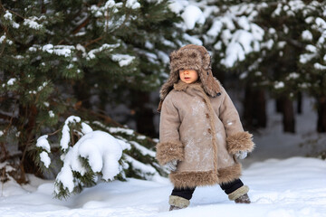 A cute boy in a fur coat and a hat with earflaps walks in winter