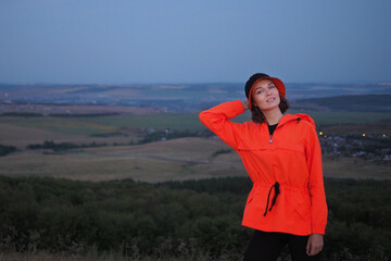 A beautiful young sports girl in a bright orange jacket stands against the backdrop of a landscape. Place for text. The concept of a healthy lifestyle, sports, tourism.