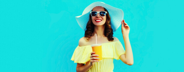 Portrait of beautiful smiling young woman drinking a fresh juice wearing a summer hat on blue background
