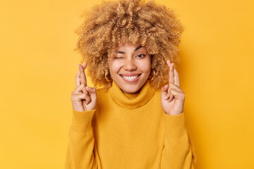Positive hopeful young woman keeps fingers crossed waits for good news winks eye smiles happily...