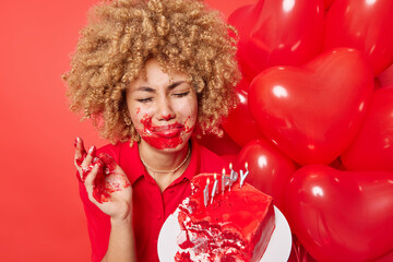 Discontent curly haired young woman upset to be lonely on Valentines Day holds cake smeared with cream bunch of heart shaped balloons imagines romantic date isolated over vivid red background
