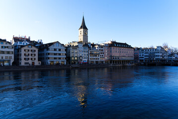 Church tower of medieval protestant church St. Peter at the old town of Zürich with river Limmat in the foreground on a sunny winter day. Photo taken February 5th, 2022, Zurich, Switzerland.
