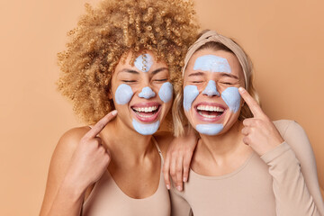 Overjoyed women apply beauty clay mask point fingers at face stand closely to each other keep eyes...