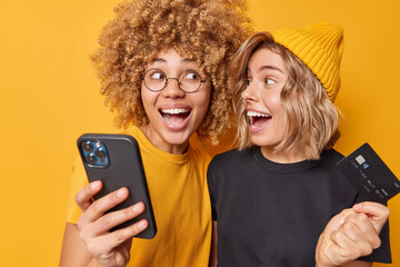 Positive glad female friends holds credit card and smartphone happy to spend money on online...