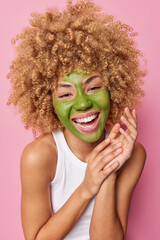 Spa beauty procedures and home skin care concept. Vertical shot of curly haired woma with green mask on face keeps hands together dressed in casual white t shirt poses against pink background